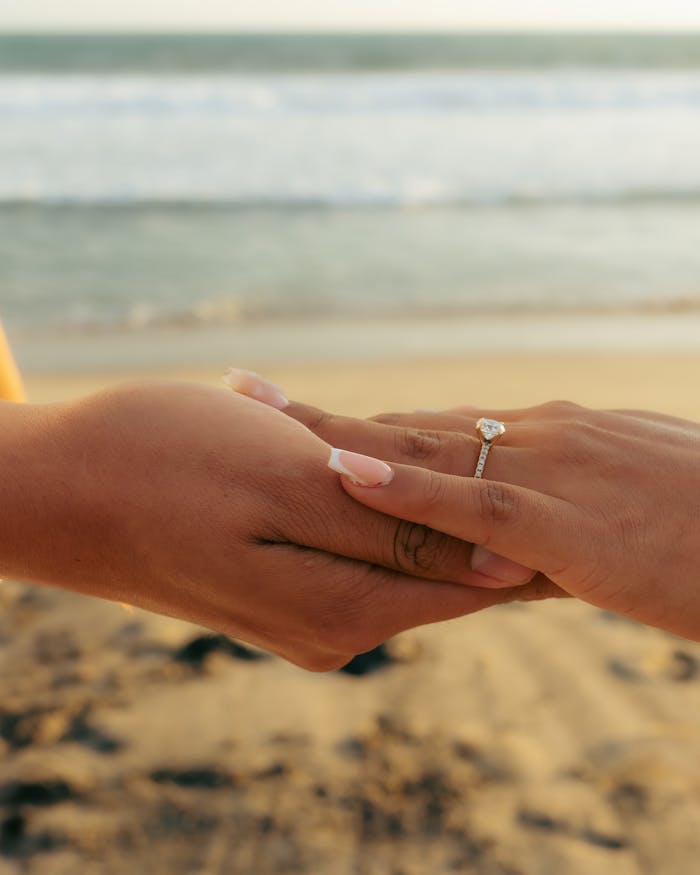 Close-up of two hands with an engagement ring against a beach backdrop.