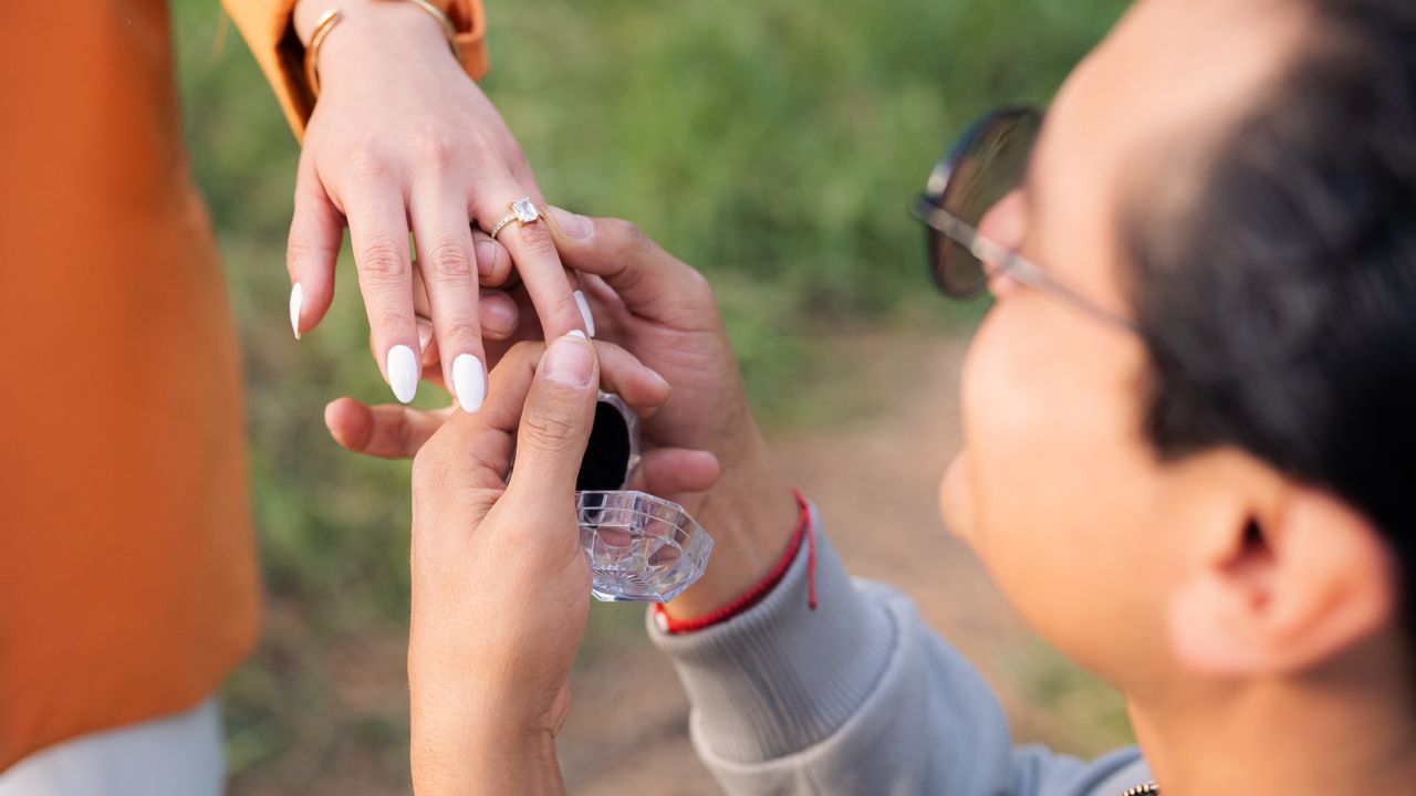 A romantic proposal scene with a man placing an engagement ring on a woman's finger outdoors.